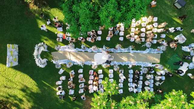 An aerial view of a large garden event, long banquet tables set out on the lawn beneath mature trees